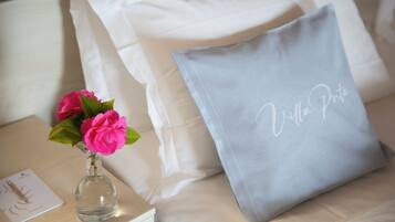 A bed with white pillows and a grey pillow with "Villa Porto" embroidered on it. A clear glass vase with pink flowers is on a bedside table.