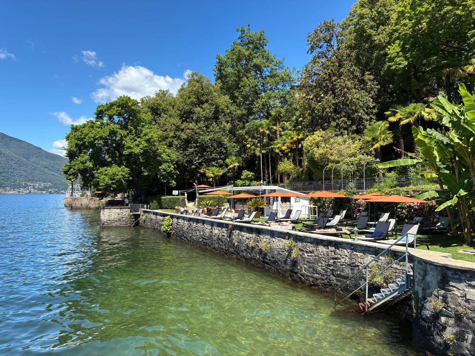 Una terraza junto al lago con sillones y sombrillas, un muro de piedra y abundante vegetación.
