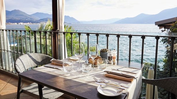 A dining table set with glasses, plates, and cutlery on a balcony overlooking a body of water with mountains in the background. - Relais Villa Porta (Luino)