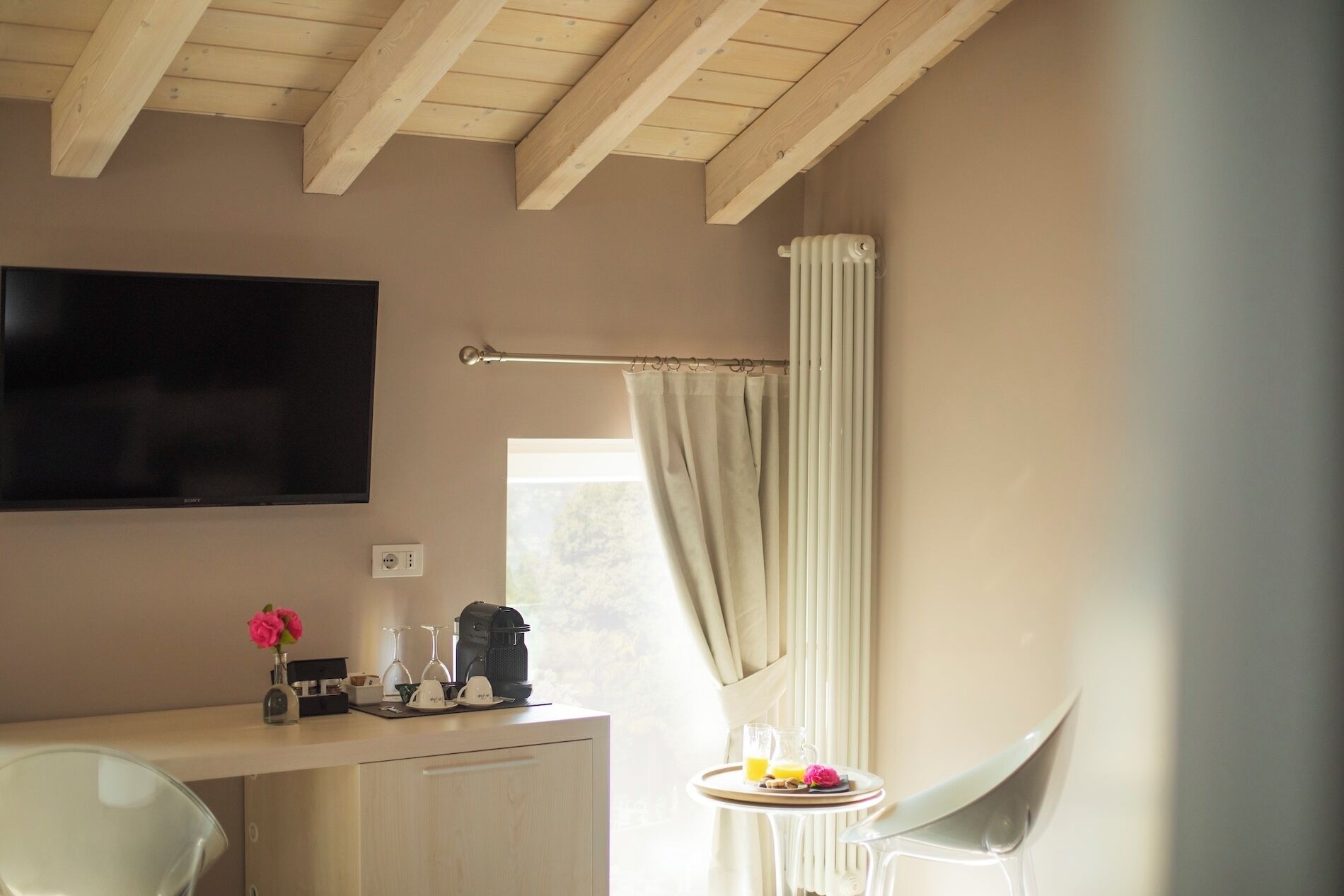 A room with a flat-screen TV mounted on the wall, a window with curtains, a small table with a tray of breakfast items, and a chair.