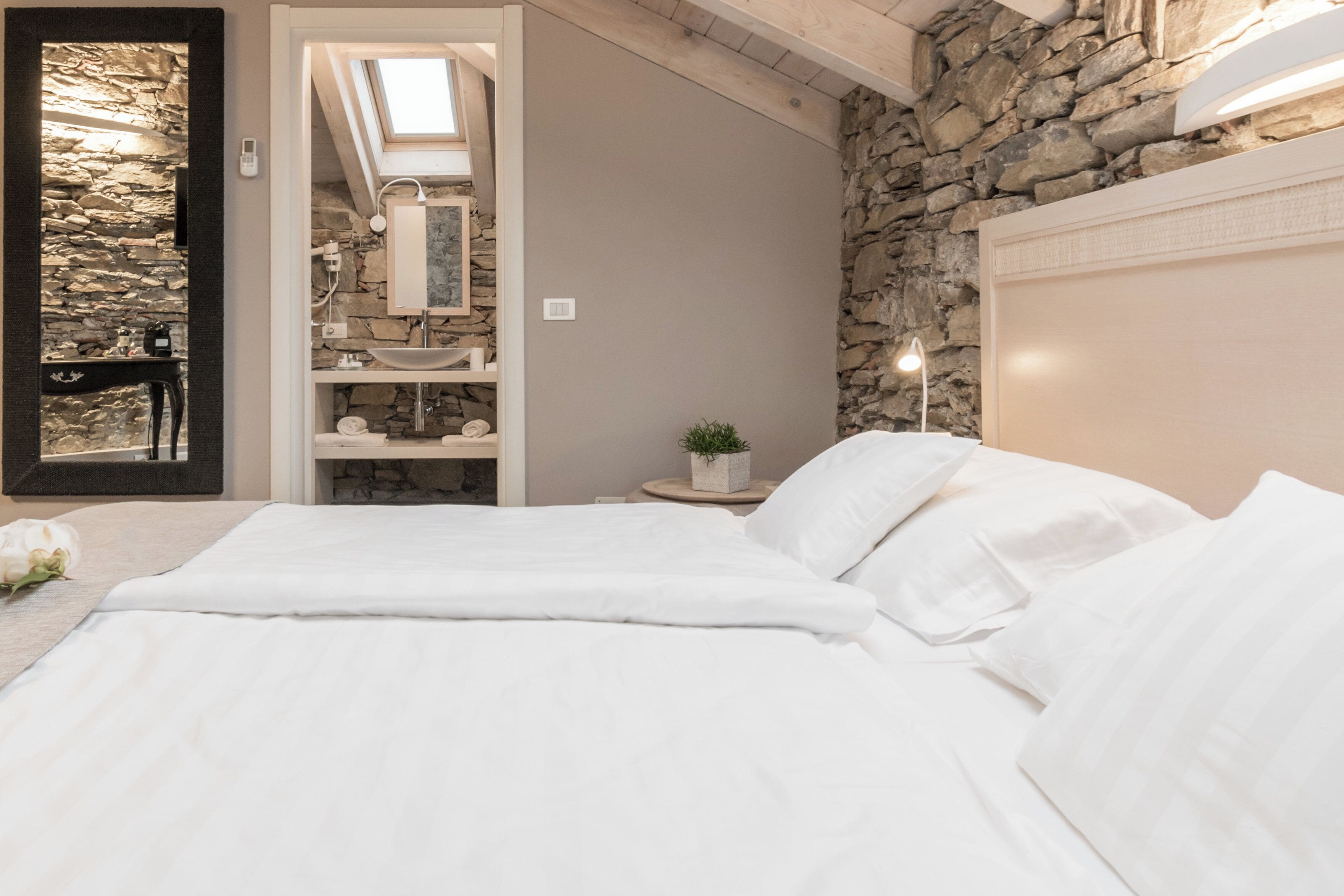 Ein Schlafzimmer mit Steinwand, Dachfenster, einem Bett mit weißen Leinen, einem Nachttisch mit Pflanze sowie einem Badezimmer mit Steinwaschbecken und Spiegel.