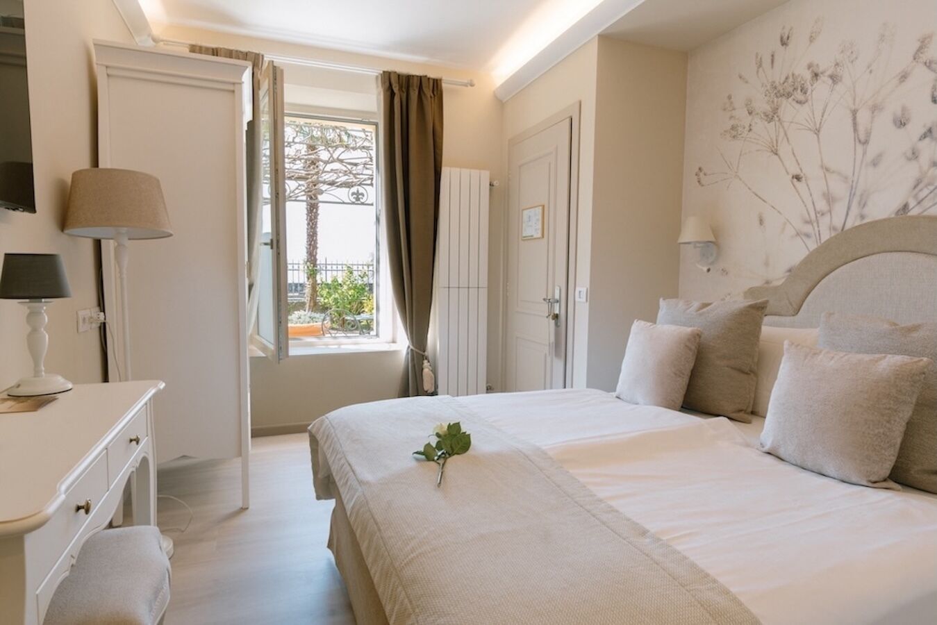 Ein Schlafzimmer mit Bett, Nachttisch, Lampe, Fenster mit Vorhängen und einer Wand mit einer Pflanze.