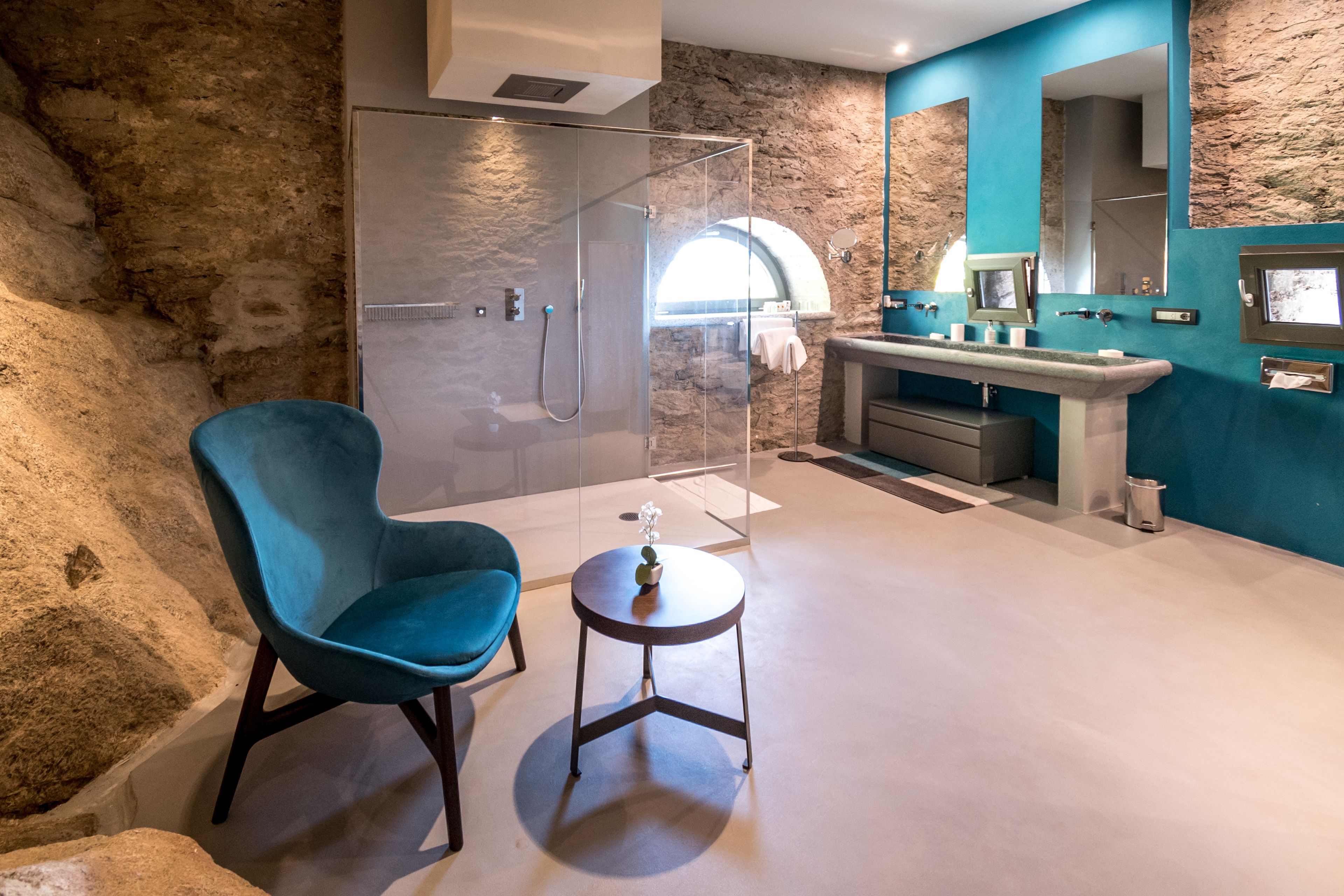 Un baño moderno con pared de piedra, cabina de ducha de cristal, una silla azul y una pequeña mesa redonda.
