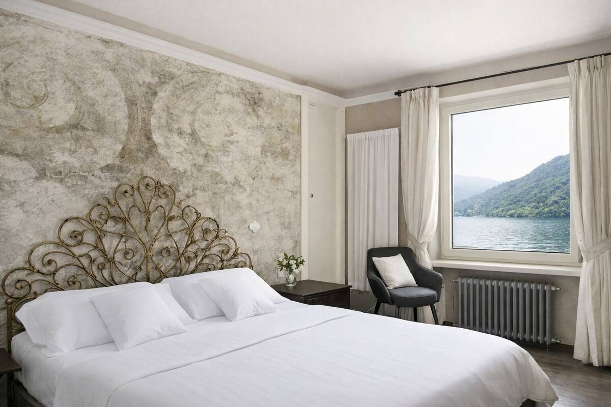 Ein Schlafzimmer mit einem großen Bett, einem Sessel, einem Nachttisch, einem Fenster mit Blick auf einen See und Berge sowie einer Heizung.