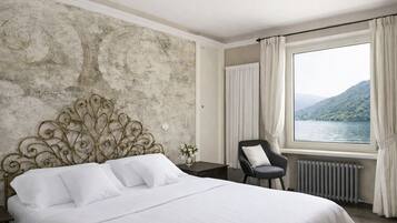 A bedroom with a large bed, a chair, a nightstand, a window with a view of a lake and mountains, and a radiator.