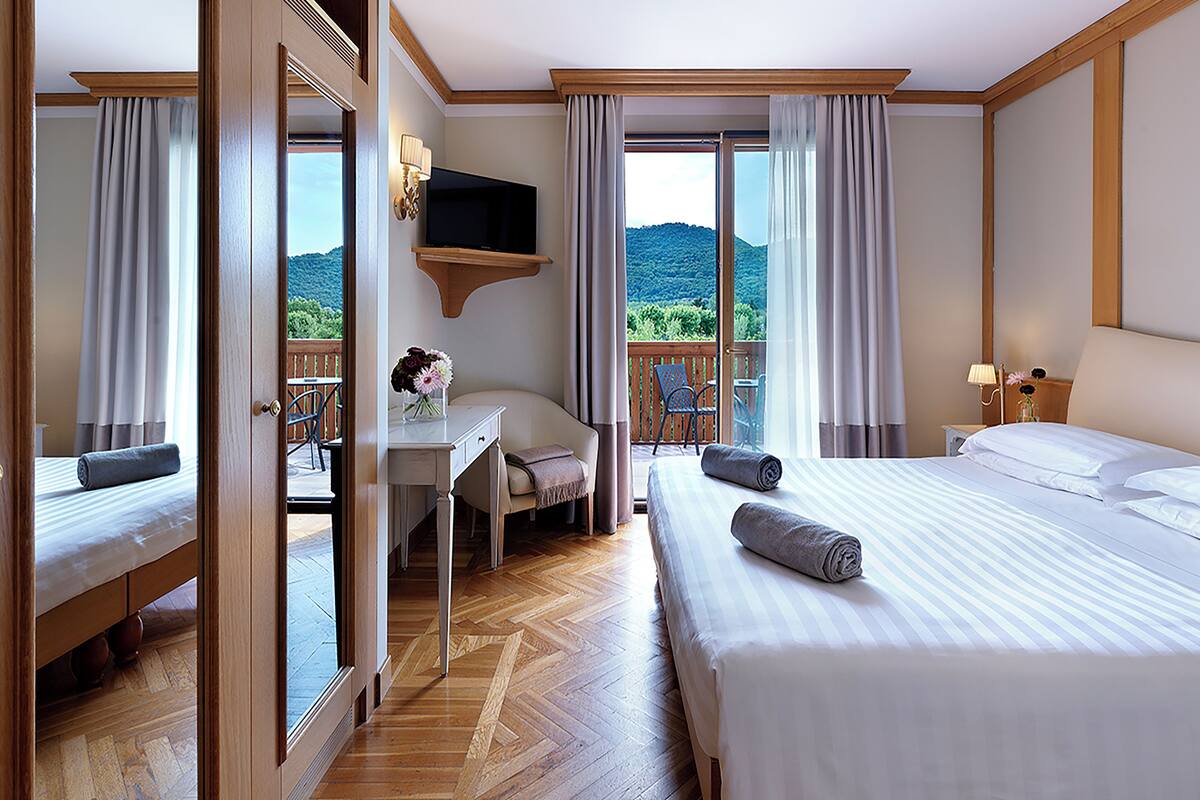 A hotel room with a bed, a desk with a chair, a TV, and a balcony with a view of the mountains.