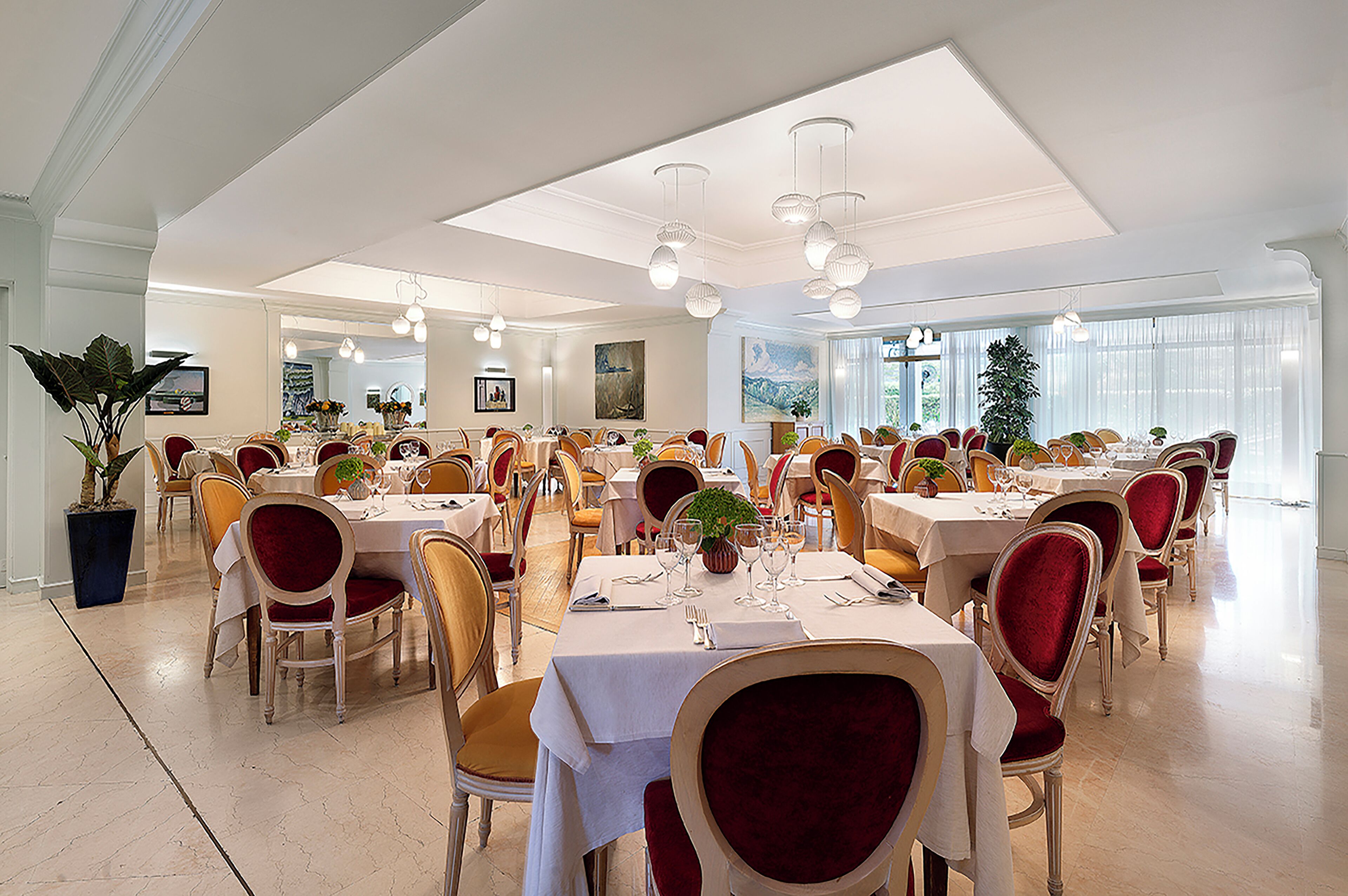 A spacious dining area with multiple tables and chairs, large windows, and a chandelier.