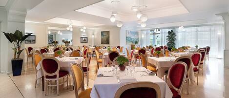 A spacious dining area with multiple tables and chairs, large windows, and a chandelier.