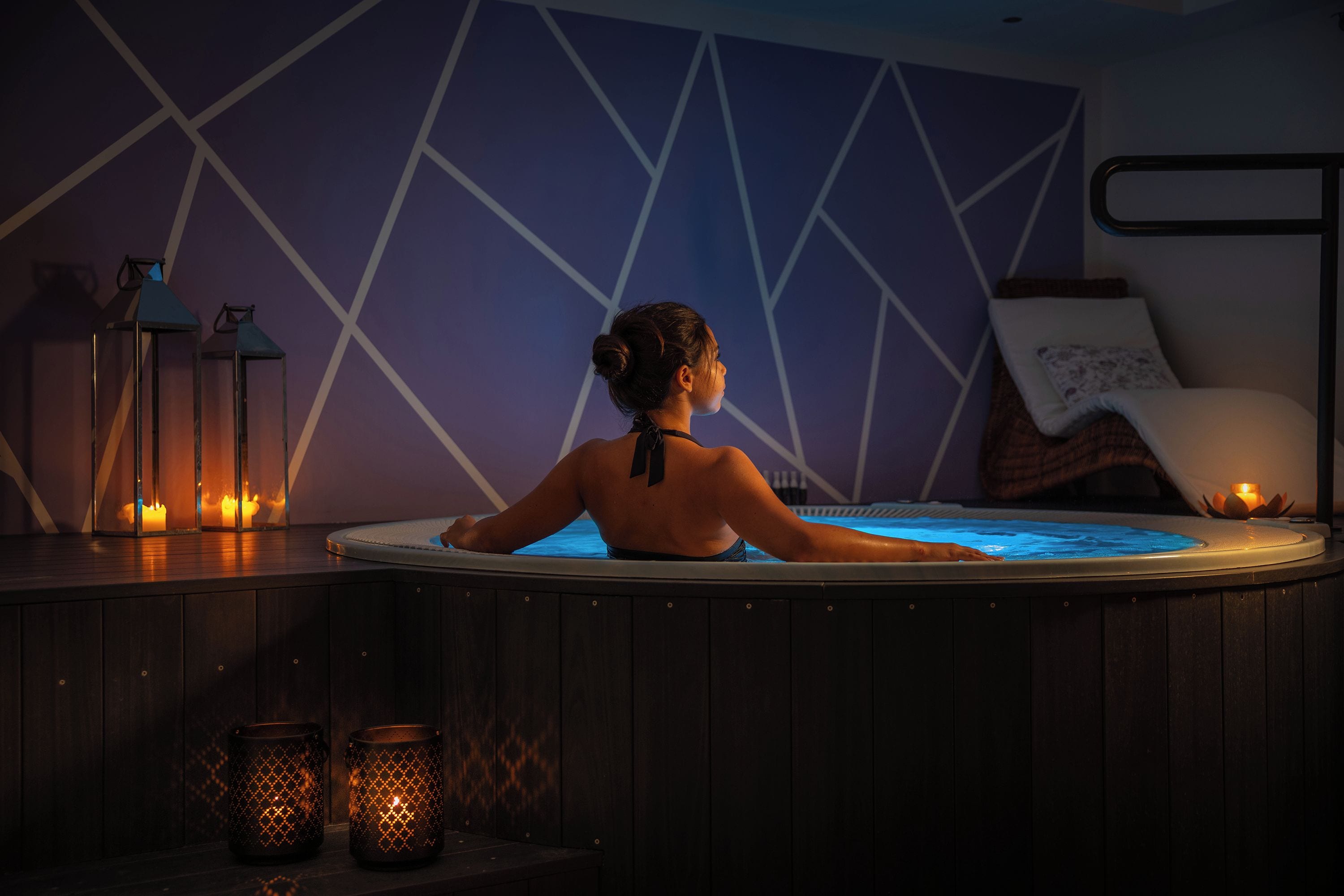 Una persona relajándose en un jacuzzi con un diseño geométrico en la pared, velas y una cama al fondo.