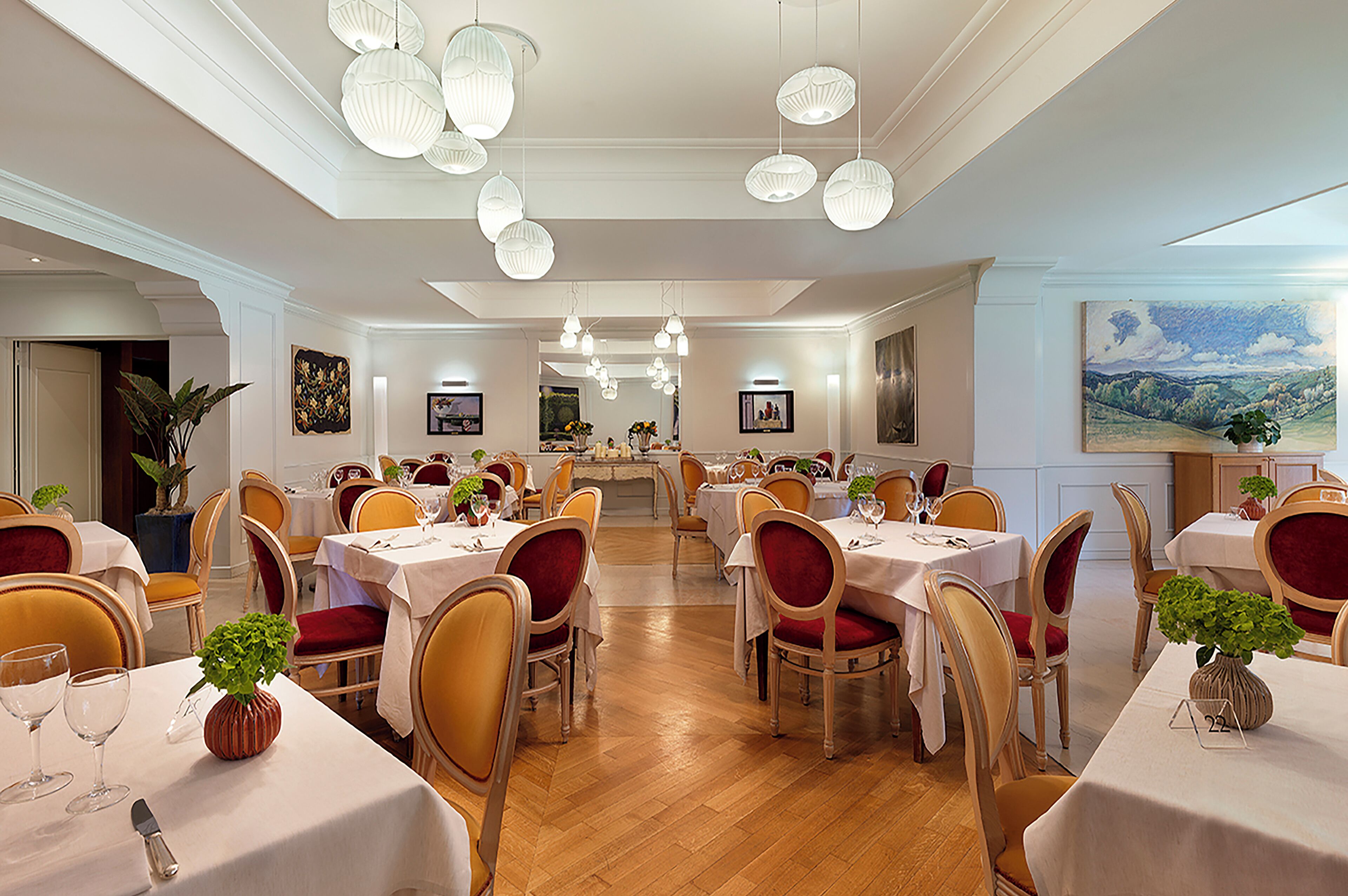 A spacious dining area with multiple tables and chairs, elegant chandeliers, and framed artwork on the walls.