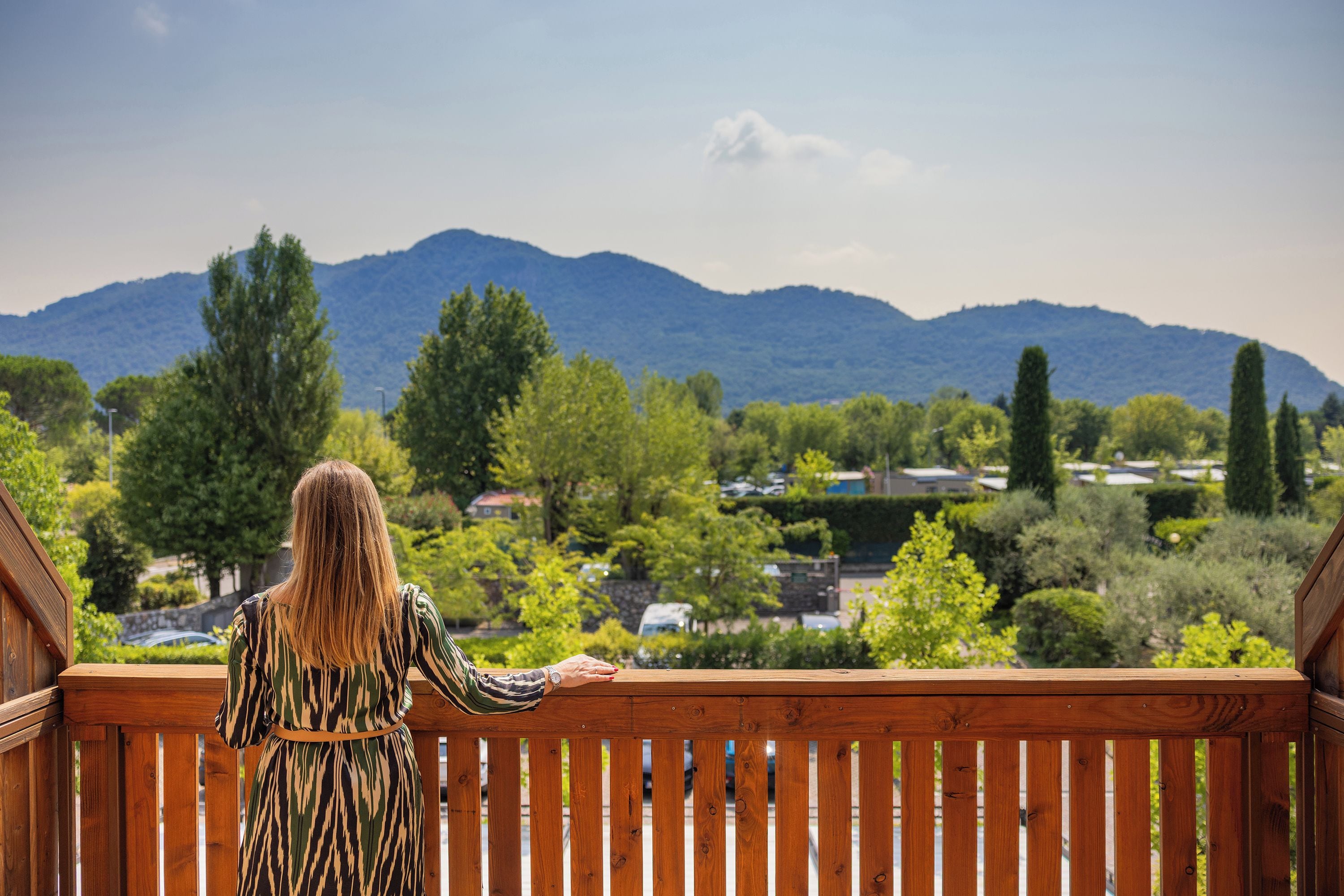 A person standing on a wooden deck overlooking a mountainous landscape with trees and houses.