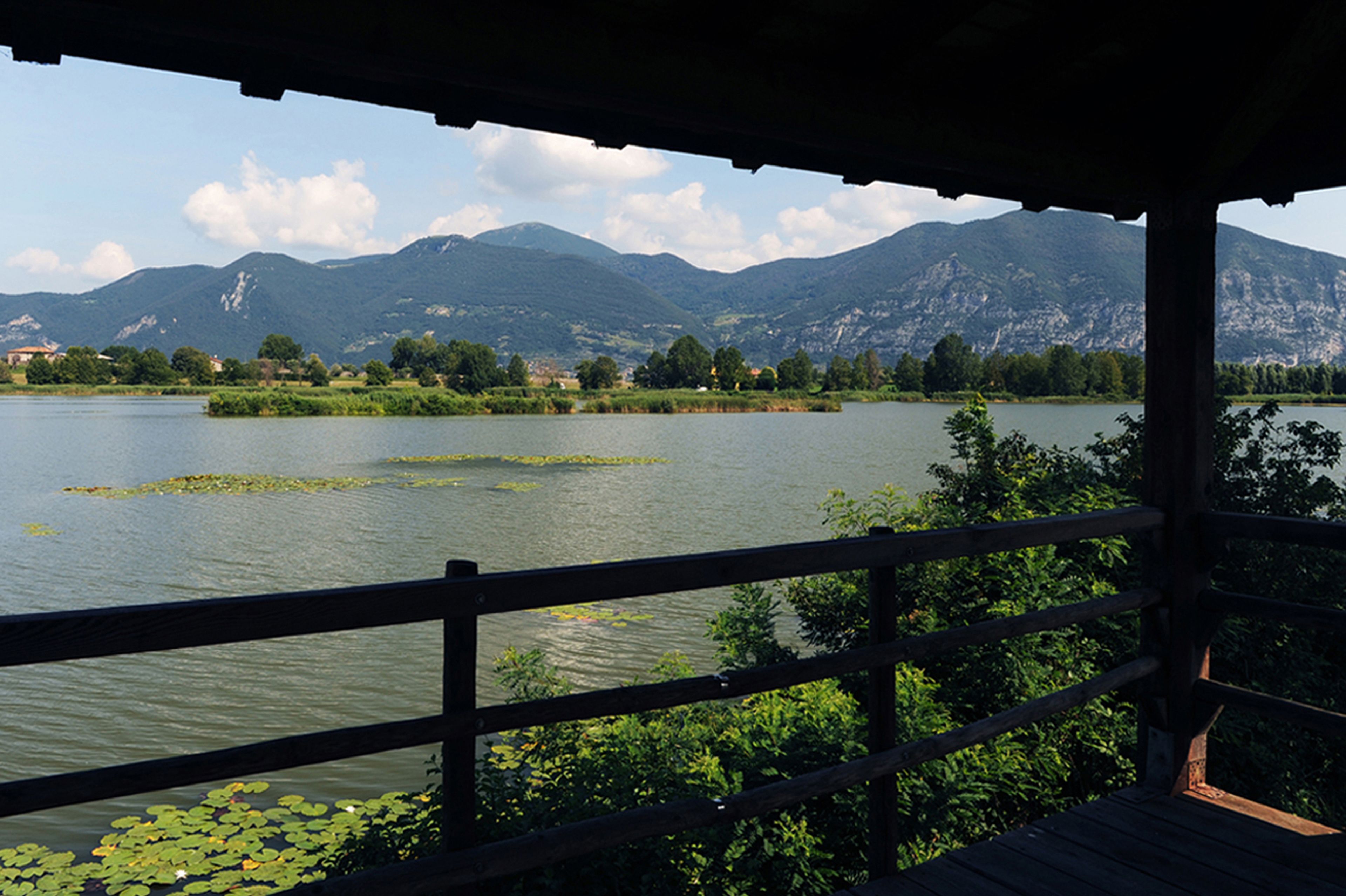 Une vue sur le lac depuis une terrasse en bois dotée d’une balustrade, entourée d’une végétation luxuriante et avec des montagnes en arrière-plan.