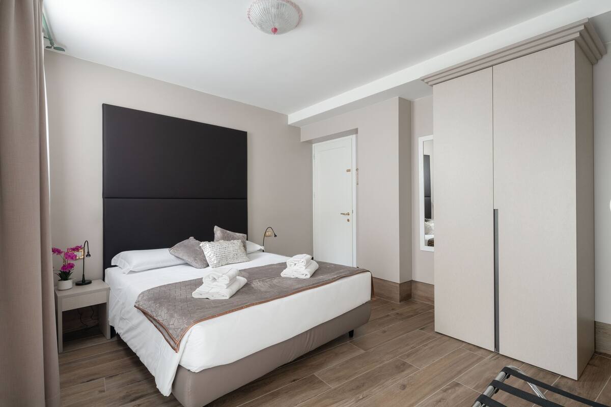 A modern bedroom with a large bed, bedside tables, a wardrobe, and a nightstand with a lamp and flowers.