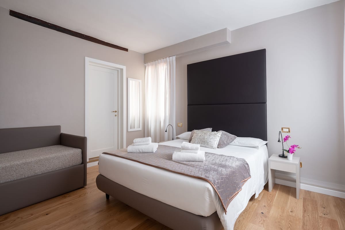 A modern bedroom with a large bed, a sofa, and a nightstand with a lamp and flowers.