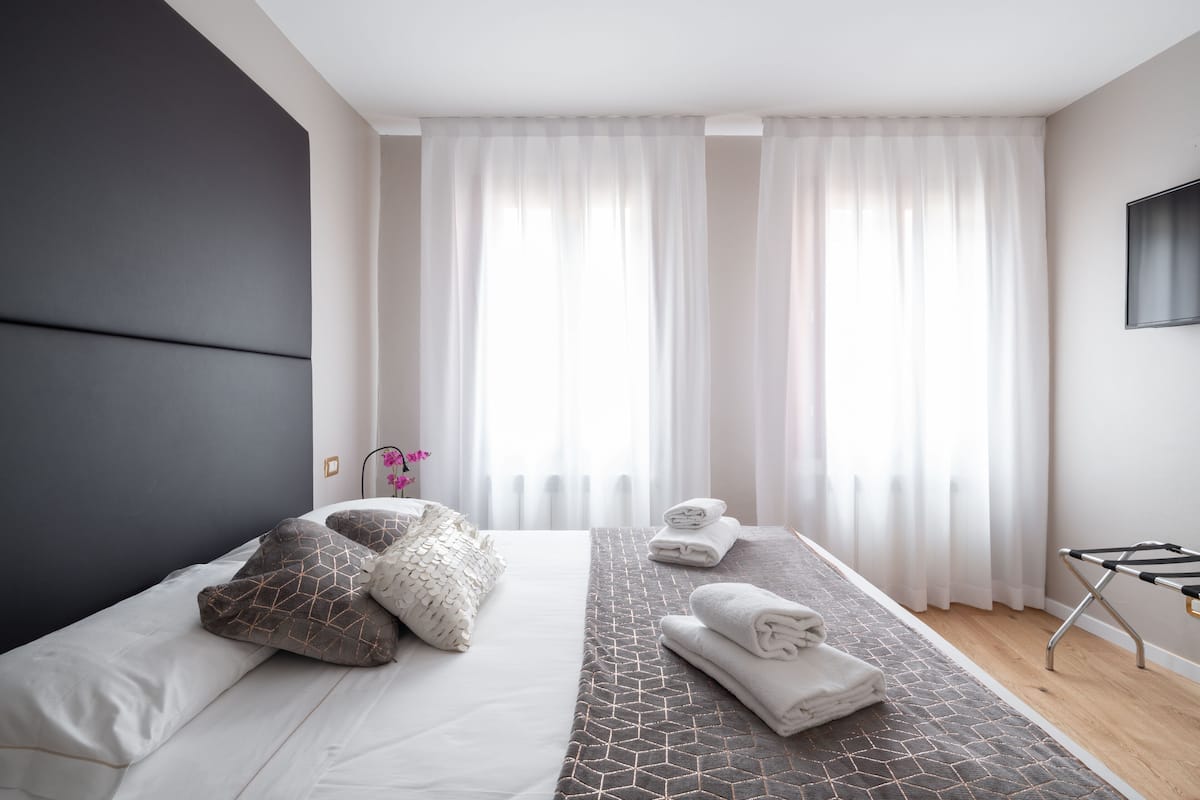 A modern hotel room with a large bed, a bedside table, a TV mounted on the wall, and a window with sheer curtains.