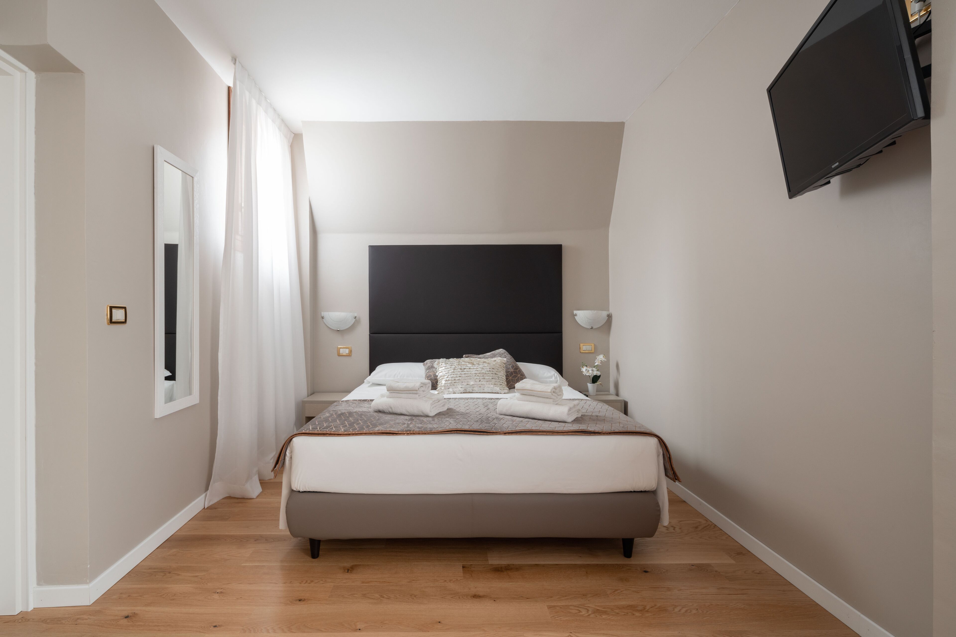 A modern bedroom with a large bed, bedside lamps, a flat-screen TV, and a window with curtains.