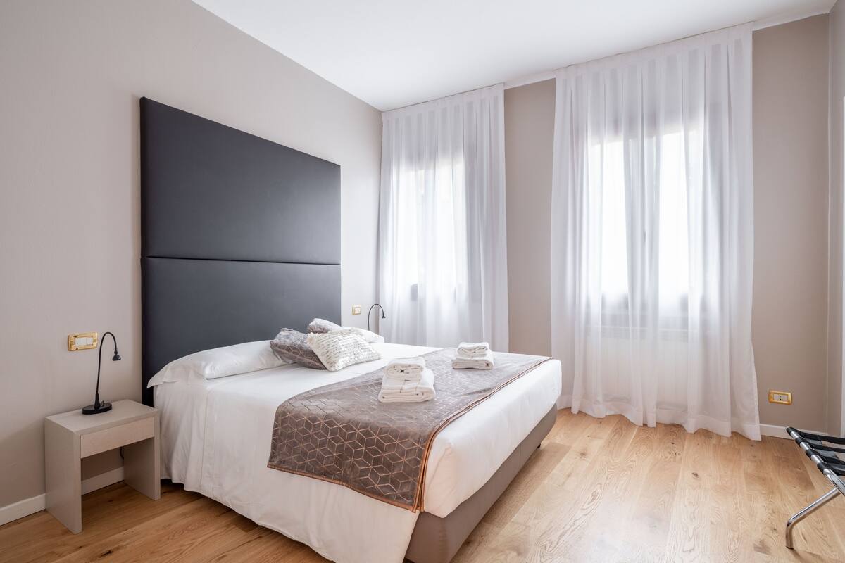A modern bedroom with a large bed, a bedside table, and a window with sheer curtains.