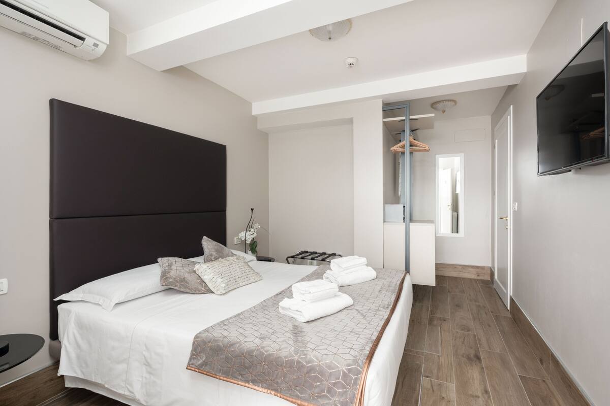 A modern hotel room with a large bed, a flat-screen TV, a walk-in closet, and a bathroom with a mirror and towel rack.