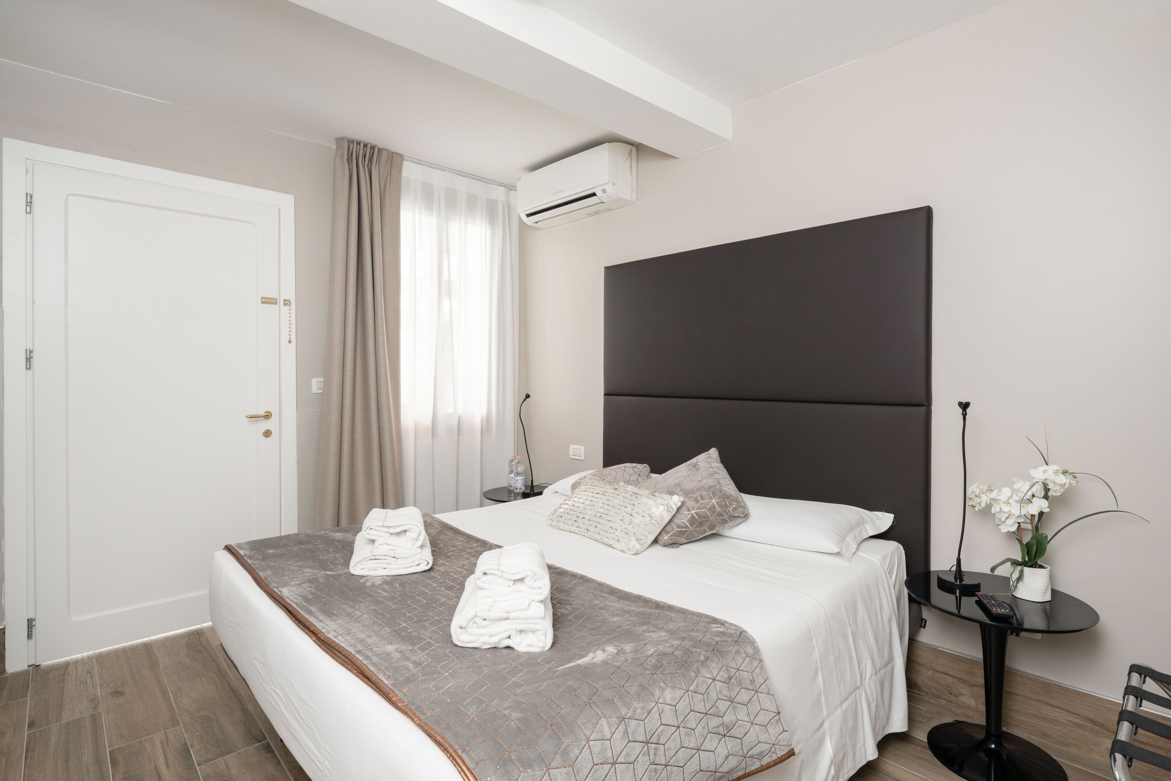Habitación de hotel moderna con una cama grande, una mesita de noche, un equipo de aire acondicionado, una pequeña mesa con un florero y una silla.