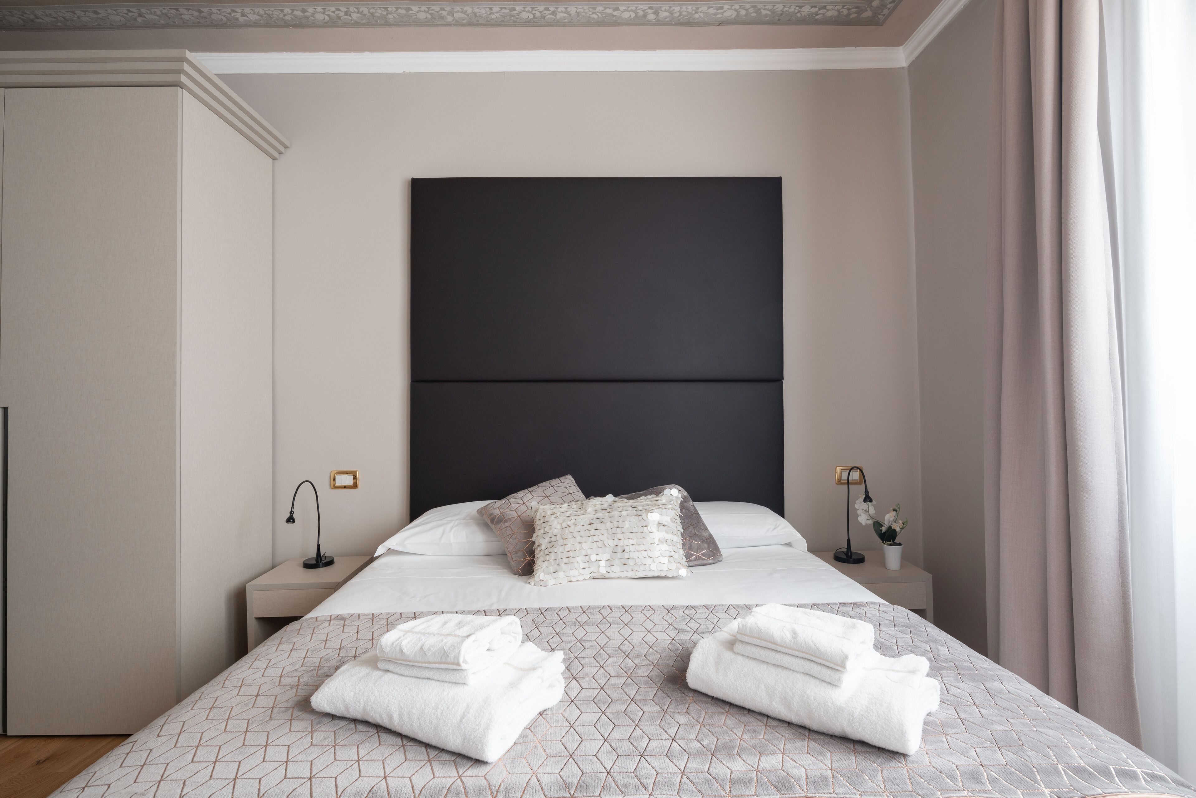 Una cama bien hecha con colcha de estampado geométrico, dos almohadas y toallas dobladas sobre un cabecero con panel oscuro.