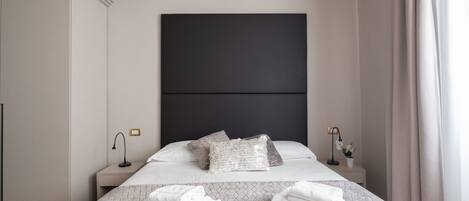 Una cama bien hecha con colcha de estampado geométrico, dos almohadas y toallas dobladas sobre un cabecero con panel oscuro.