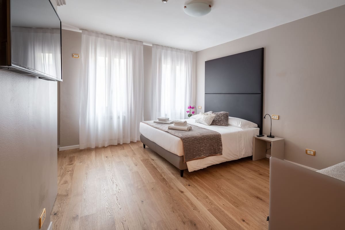 A modern bedroom with a large bed, a bedside table, a flat-screen TV, and a large window with sheer curtains.