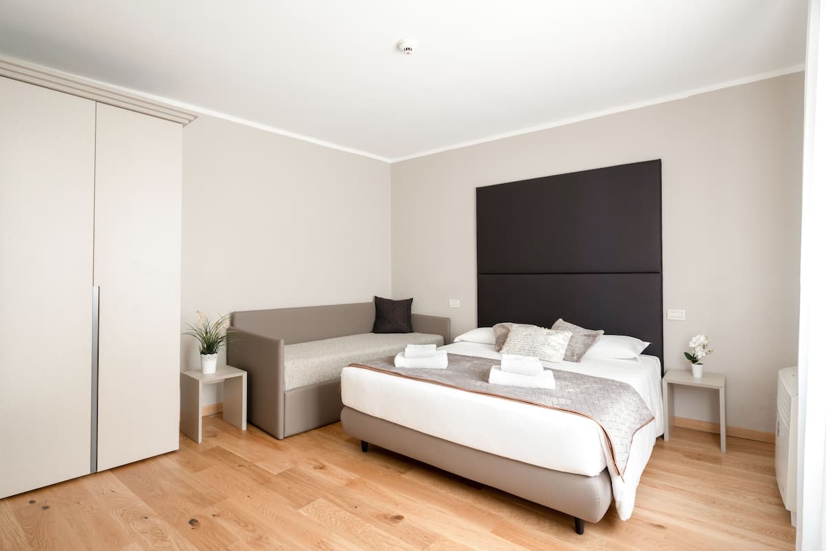A modern bedroom with a large bed, a sofa bed, a small bedside table with a plant, and a nightstand with a lamp.