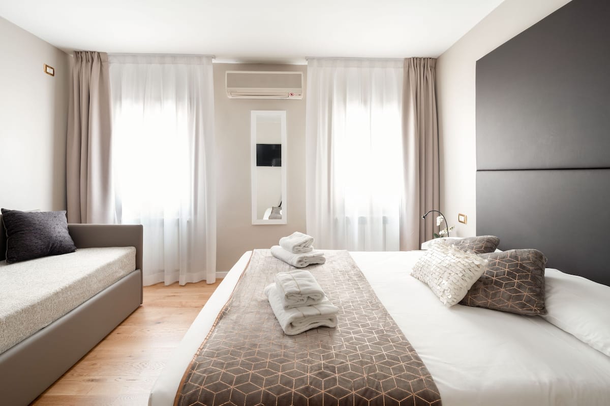 A hotel room with a bed, a sofa, and a large window with curtains. There is a wall-mounted air conditioner and a mirror above the bed.