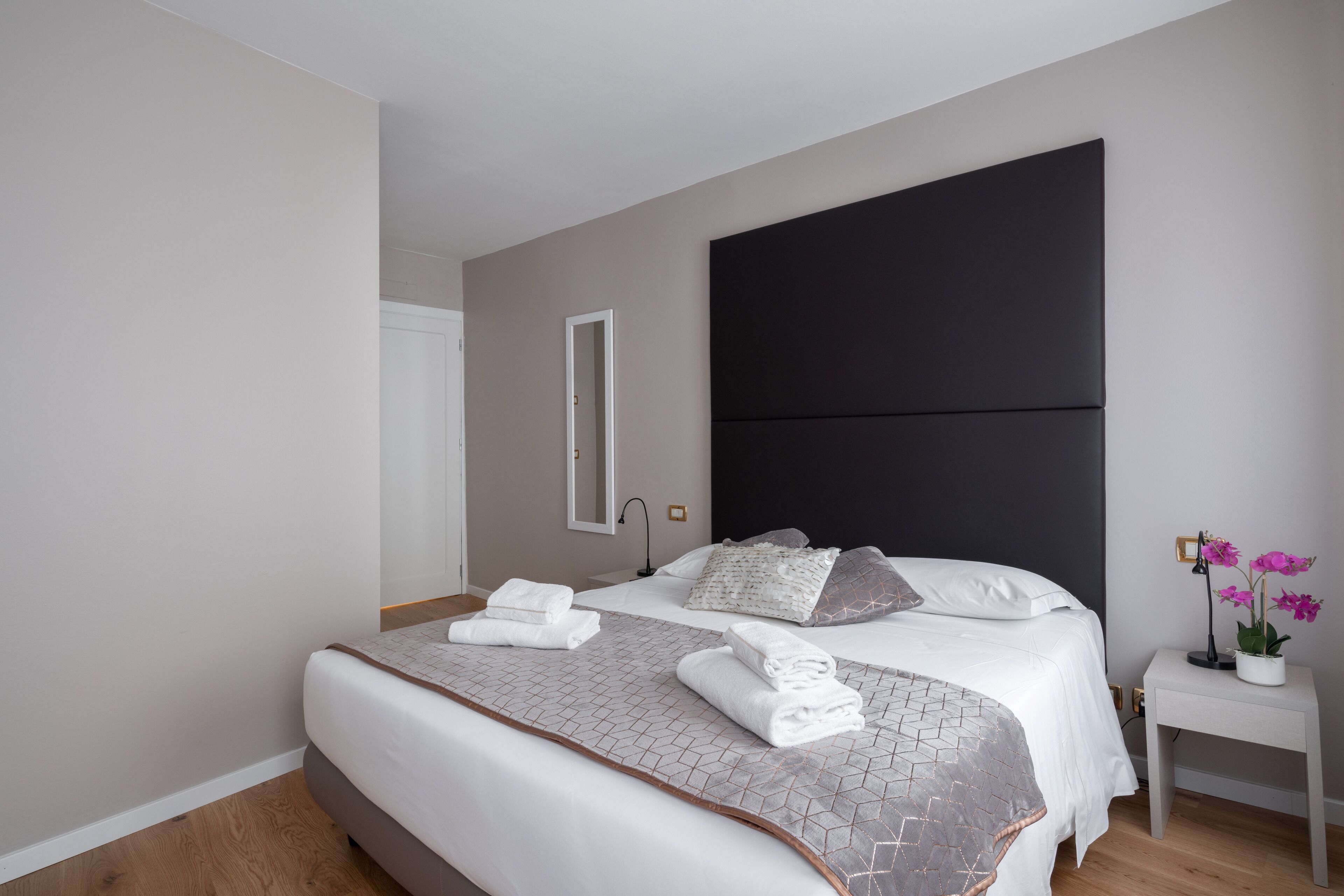 A modern bedroom with a large bed, a dark headboard, a bedside table with a lamp, and a potted plant.