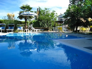 Piscina con sillas de descanso y sombrillas, rodeada de vegetación y un edificio.