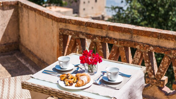 Una mesa con preparación para desayuno que incluye croissants y un plato de comida, dos tazas y un florero con flores en un balcón con vista a un paisaje urbano.