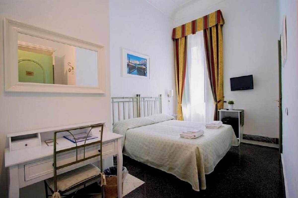 A hotel room with a bed, a desk with a chair, a TV, and a window with curtains.