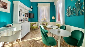 A room with teal walls, white furniture, and a tiled floor. There is a television mounted on the wall, a small table with chairs, and a bookshelf with various items. - Hotel Posta (Syracuse)