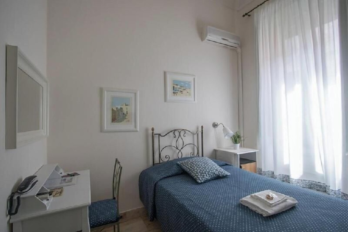 A bedroom with a bed, a desk, a chair, a small table, and two framed pictures on the wall.
