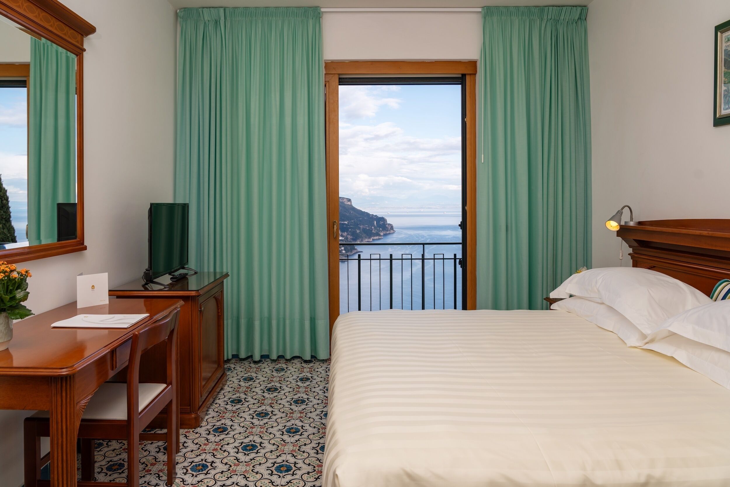 Een hotelkamer met een bed, een bureau, een stoel, een televisie en uitzicht op zee vanaf een balkon.