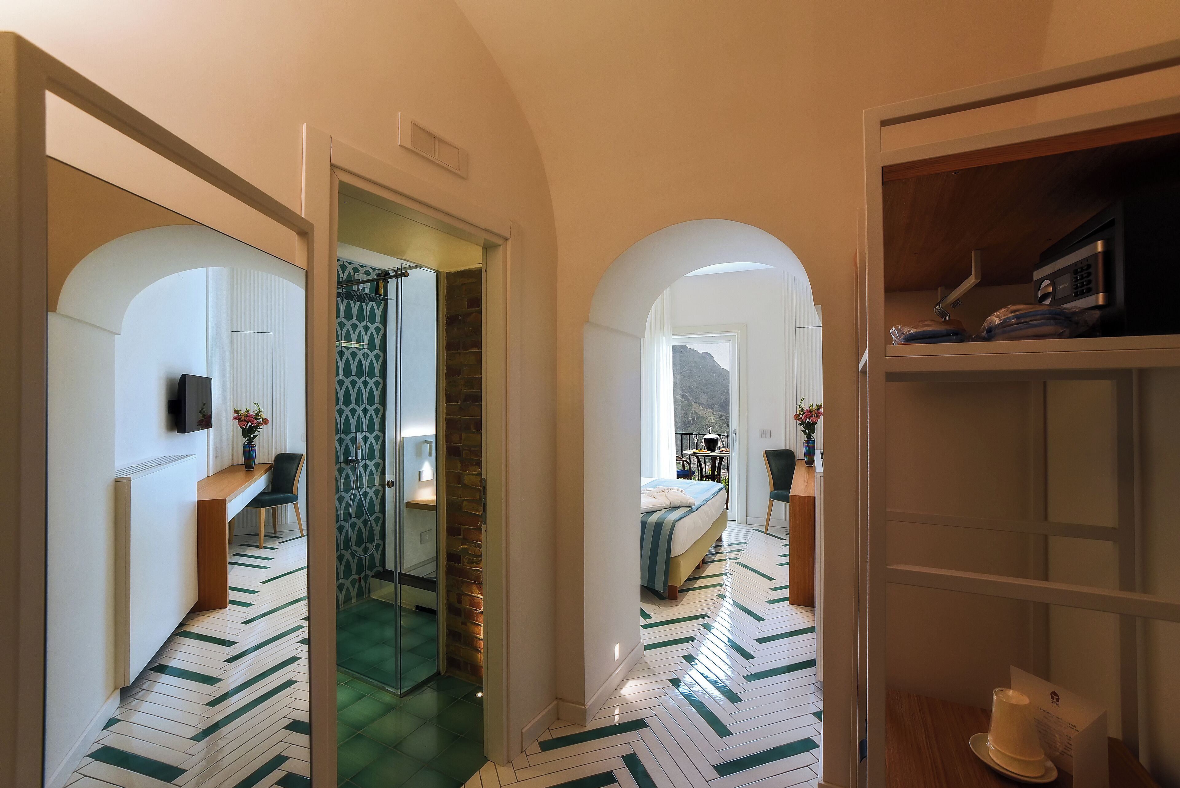 Un pasillo que conduce a un dormitorio con una cama, un escritorio y una silla. Hay una puerta de vidrio y una vista de la montaña.