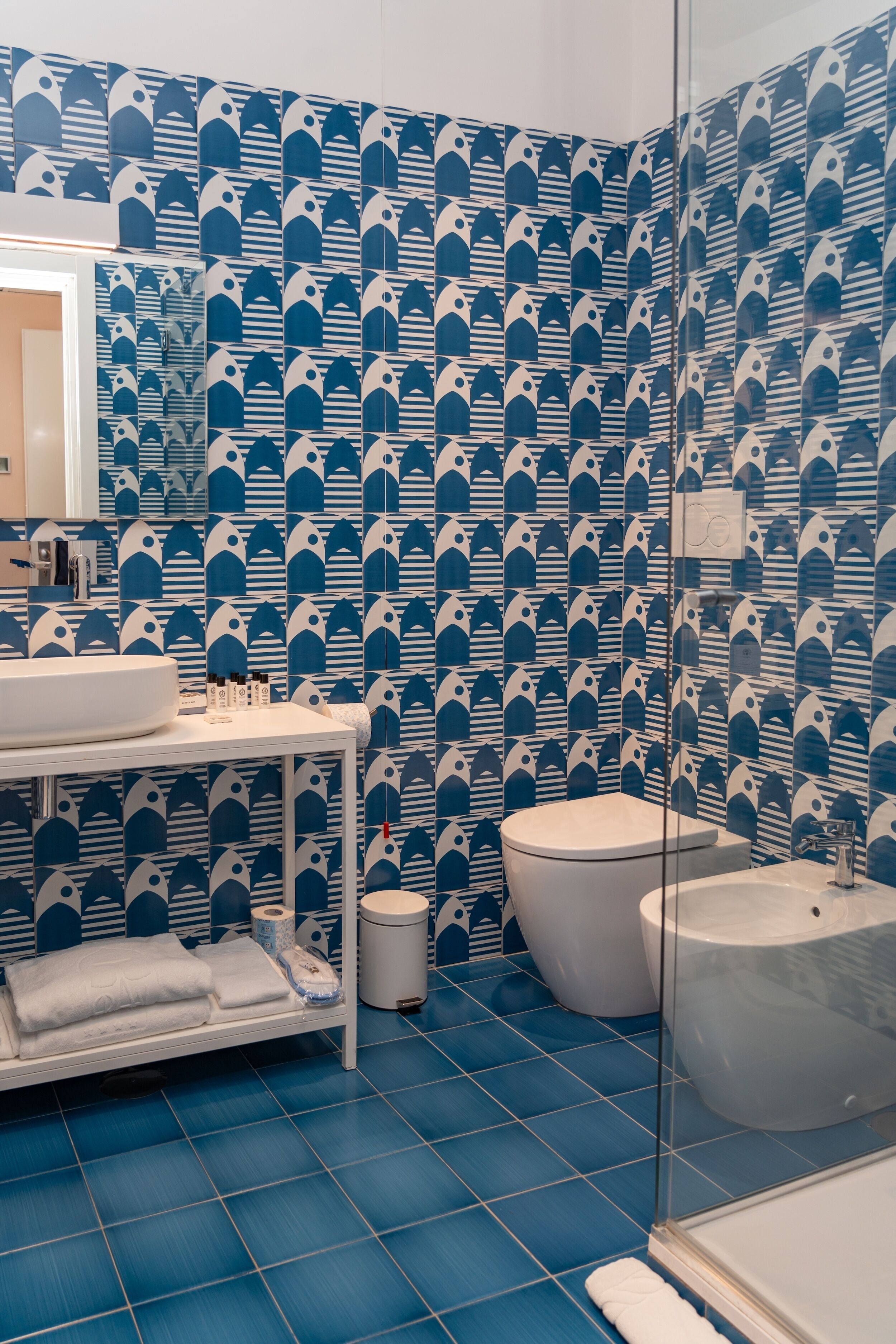 Un baño con piso de baldosas azules, un lavamanos blanco, un inodoro y una cabina de ducha con mampara de vidrio.