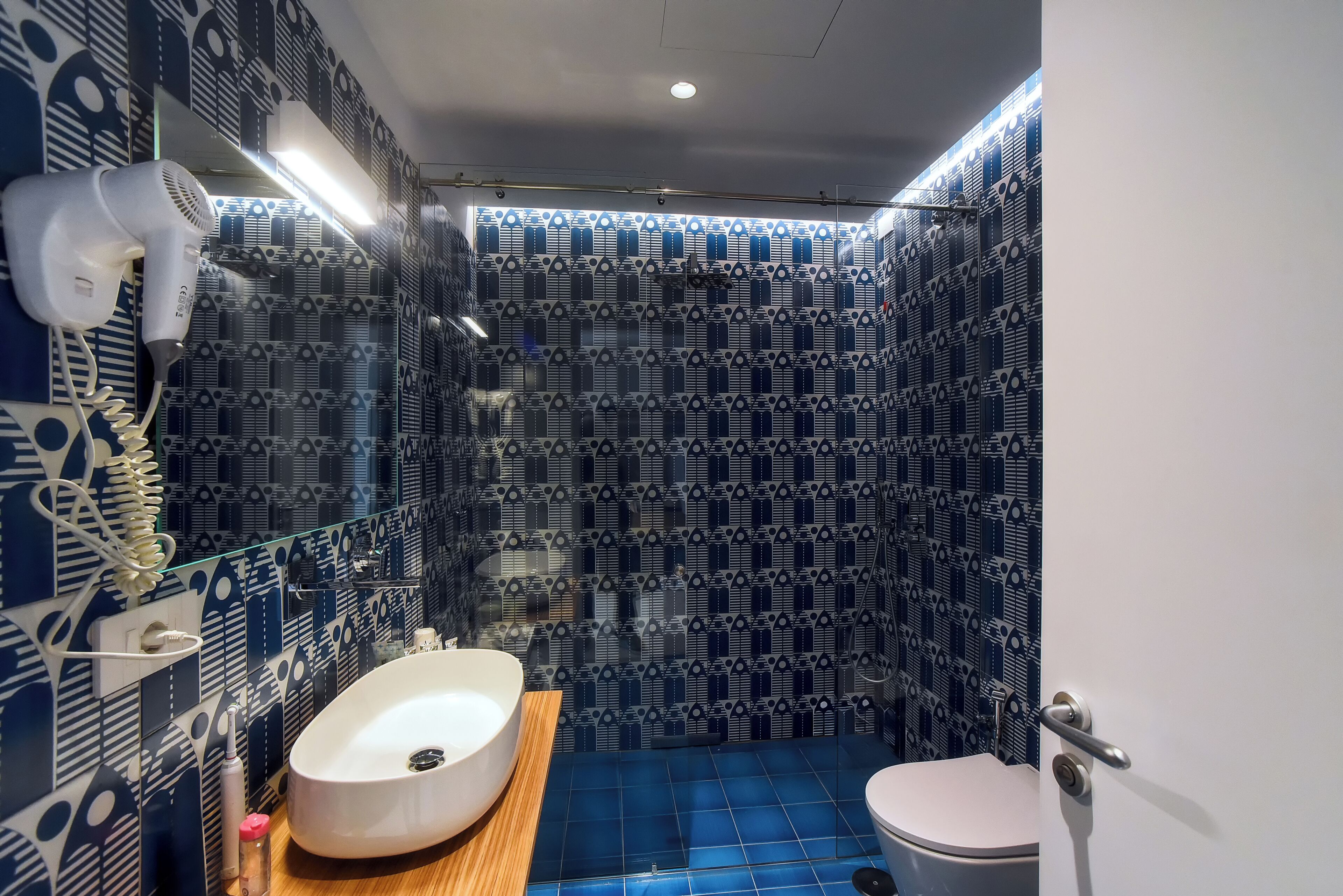 Une salle de bain avec un mur recouvert de carreaux à motifs, un lavabo blanc, un sol carrelé de bleu et un sèche-cheveux fixé au mur.