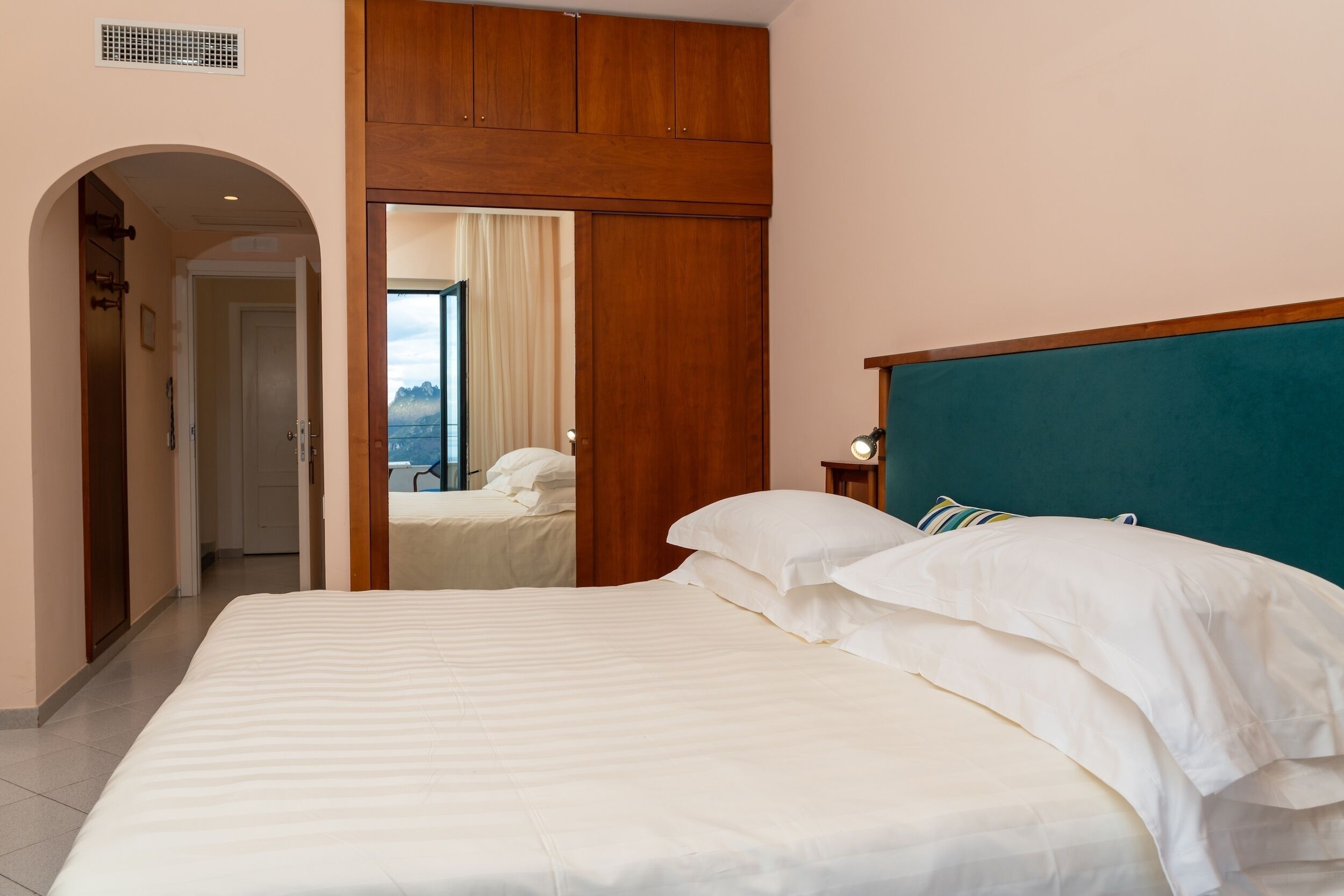 Camera d'albergo con un letto grande, testiera in legno e vista sulle montagne attraverso una porta aperta.