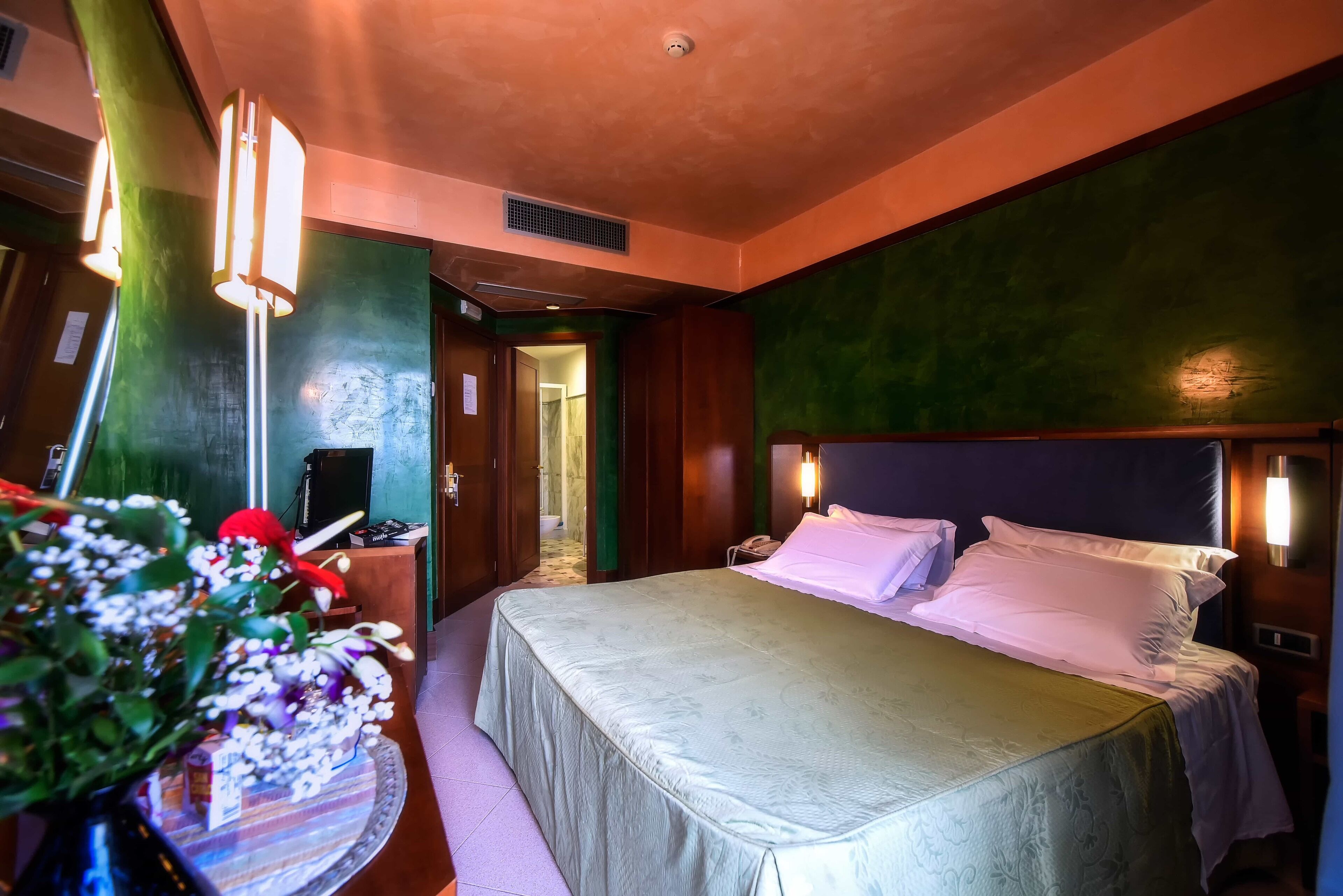 Une chambre d’hôtel avec un lit, des lampes de chevet, un bureau avec un ordinateur et un vase rempli de fleurs.