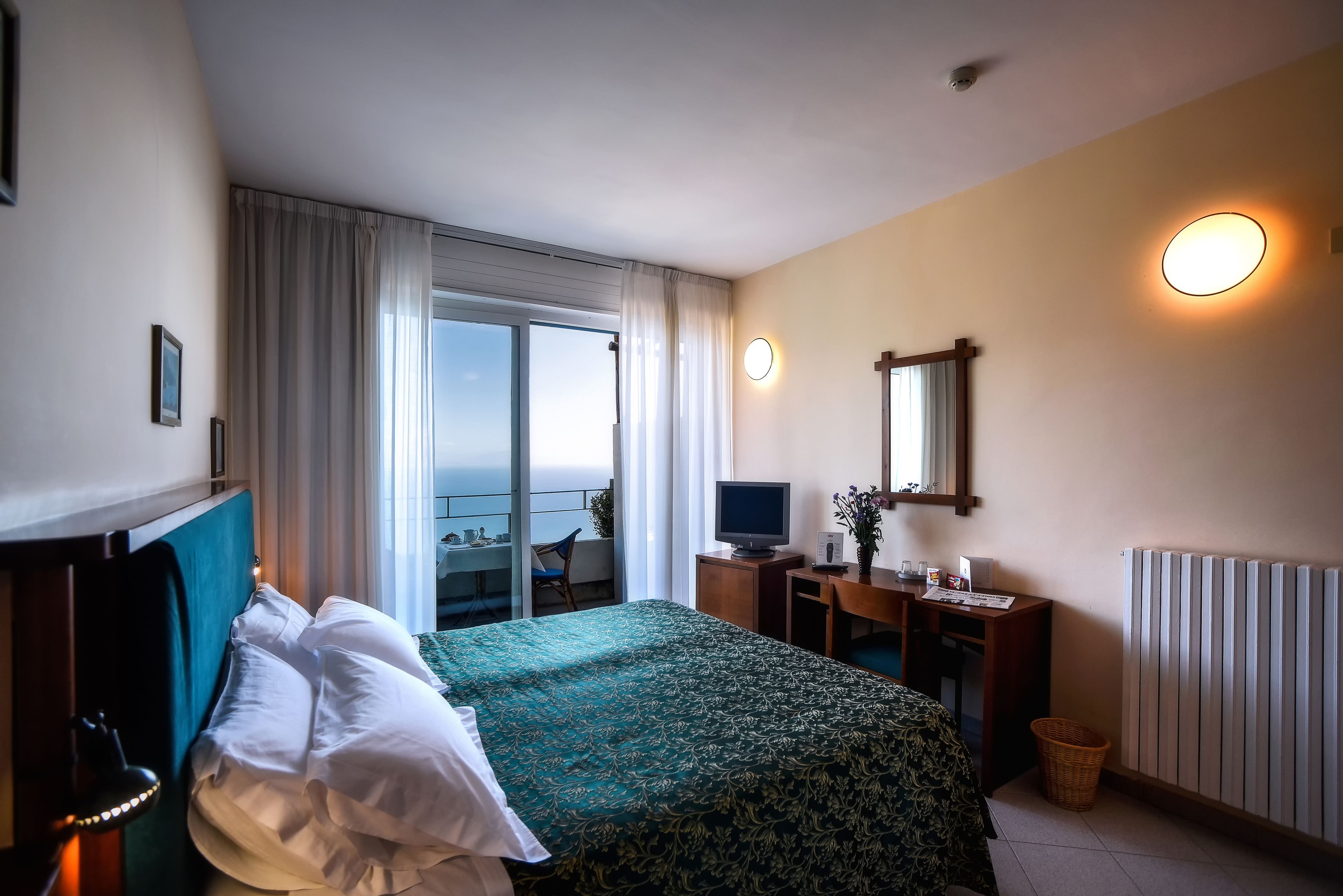 Una habitación de hotel con cama, escritorio, televisor, ventana con cortinas y vista al mar.