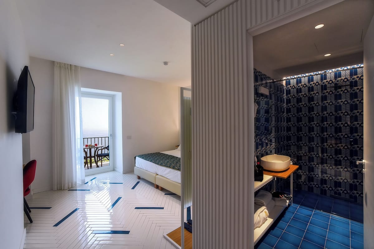 A hotel room with a bed, a bathroom with a blue tiled floor, a sink, and a mirror. There is a balcony with a table and chairs visible through the window.