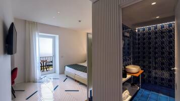 Hotelkamer met een bed, een badkamer met een blauwe betegelde vloer, een wastafel en een spiegel. Door het raam is een balkon zichtbaar met een tafel en stoelen.