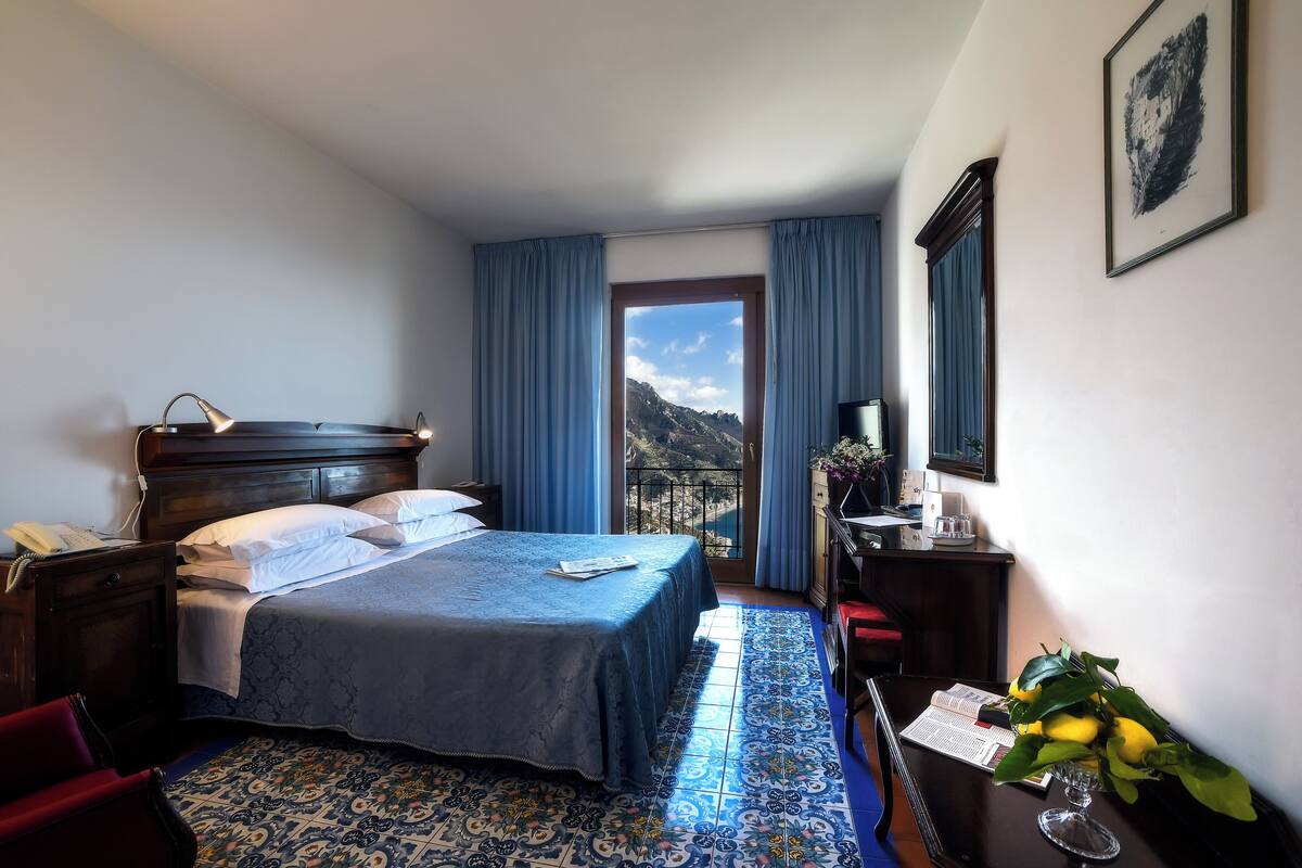 A hotel room with a bed, a desk, a chair, a window with curtains, and a view of the mountains.