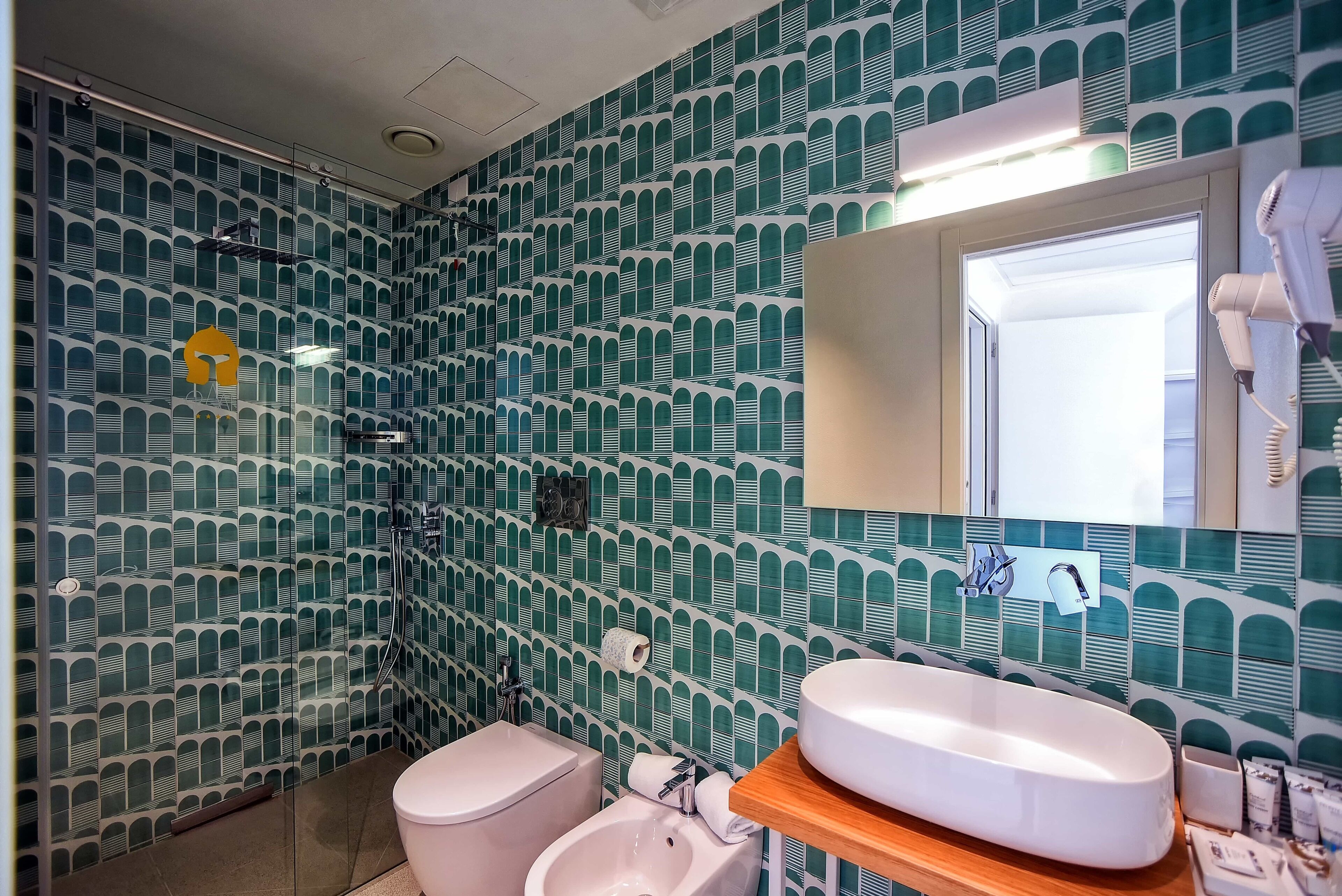 A bathroom with a patterned tile wall, a toilet, a bidet, a sink, and a mirror.