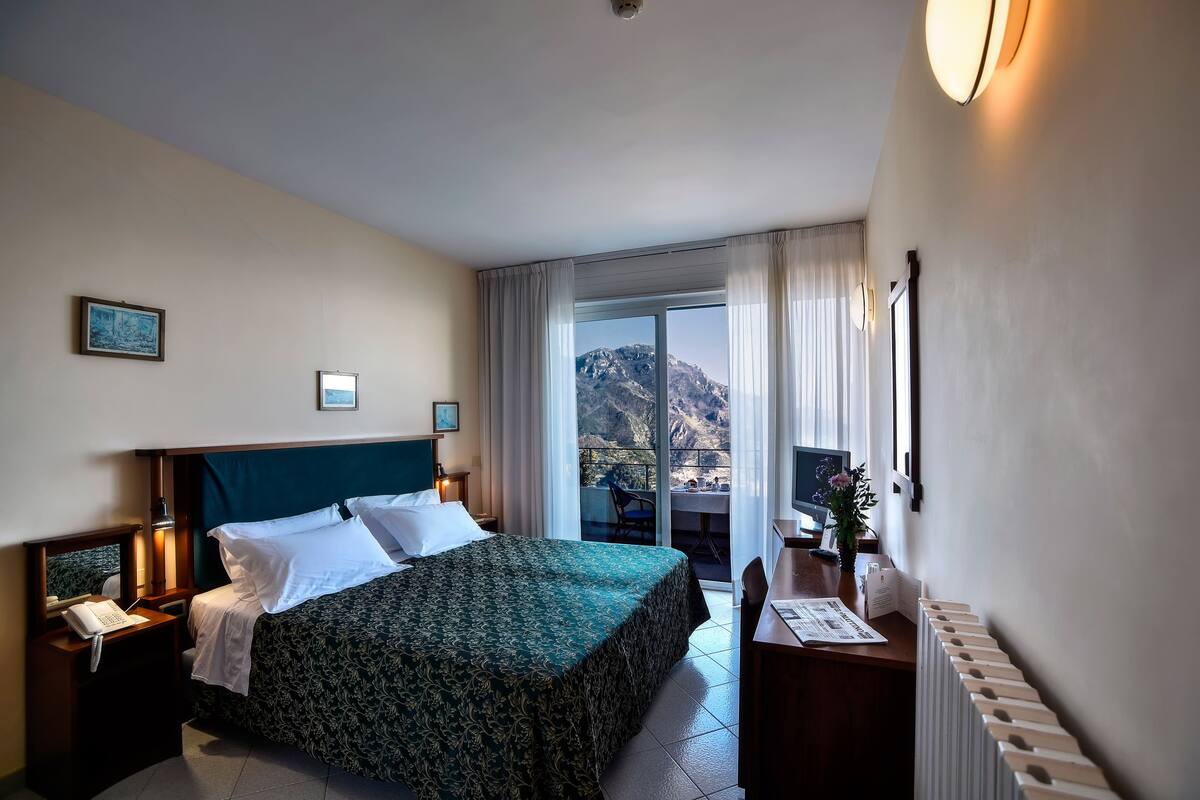 A hotel room with a bed, a desk, a chair, a telephone, and a window with a mountain view.
