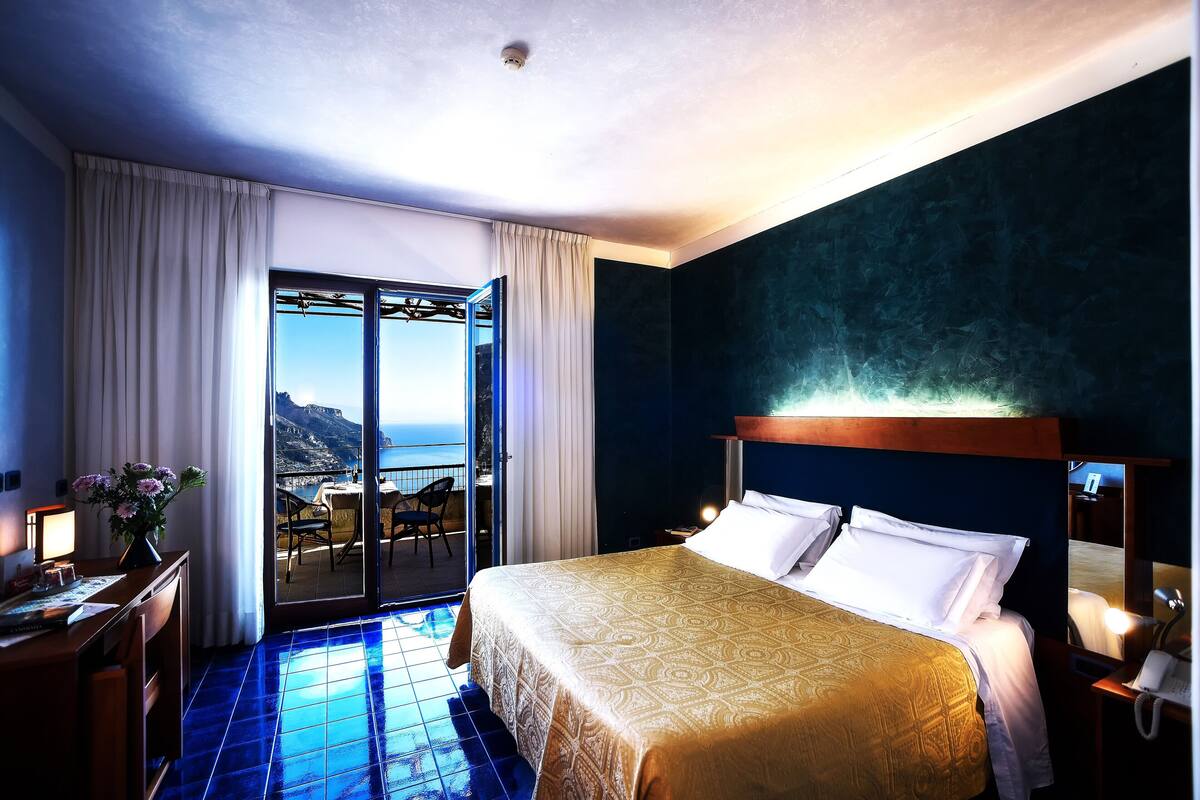 A hotel room with a bed, a desk with a lamp, a vase with flowers, and a view of the sea through a sliding door.