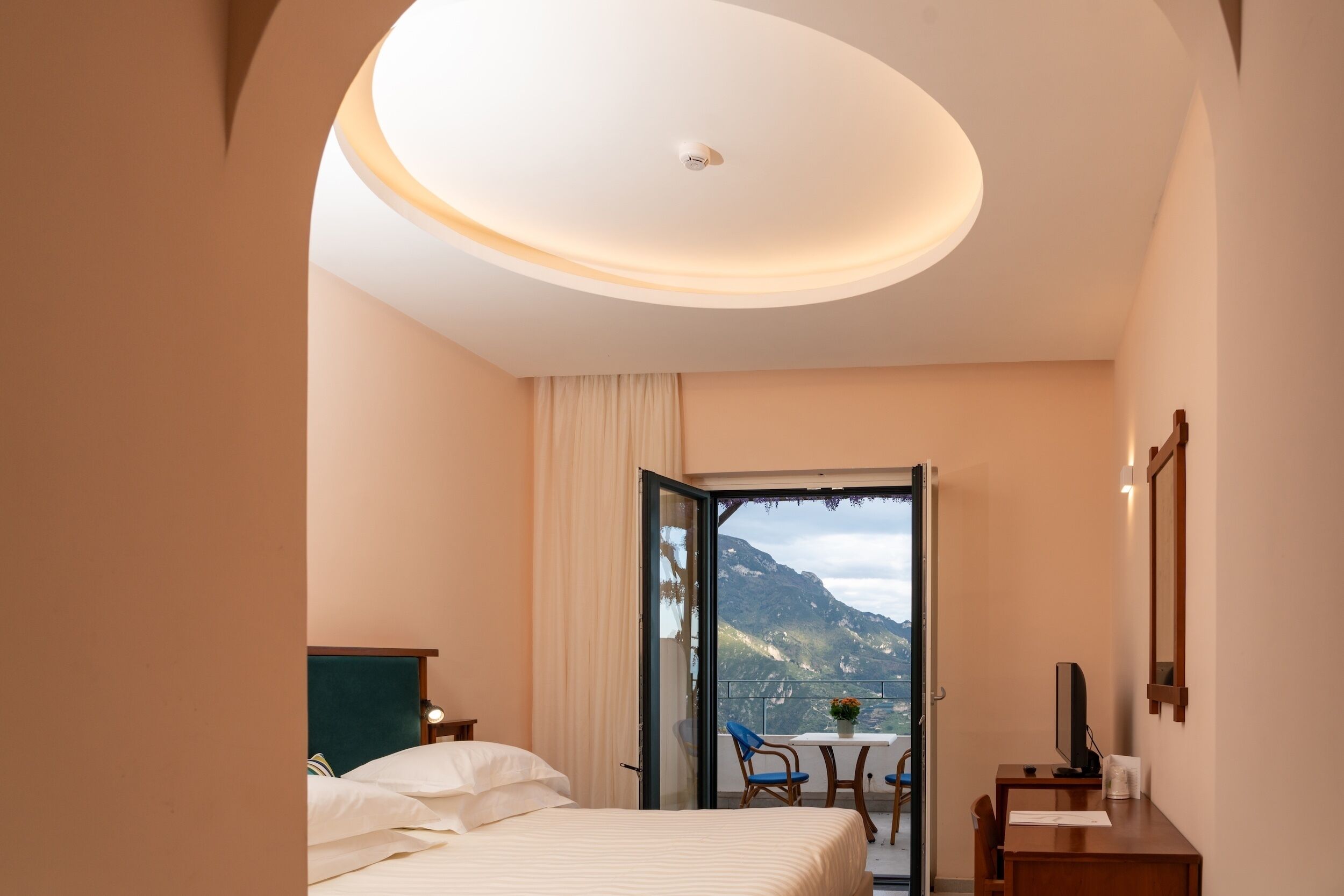 A hotel room with a bed, a desk, and a view of mountains through an open door.