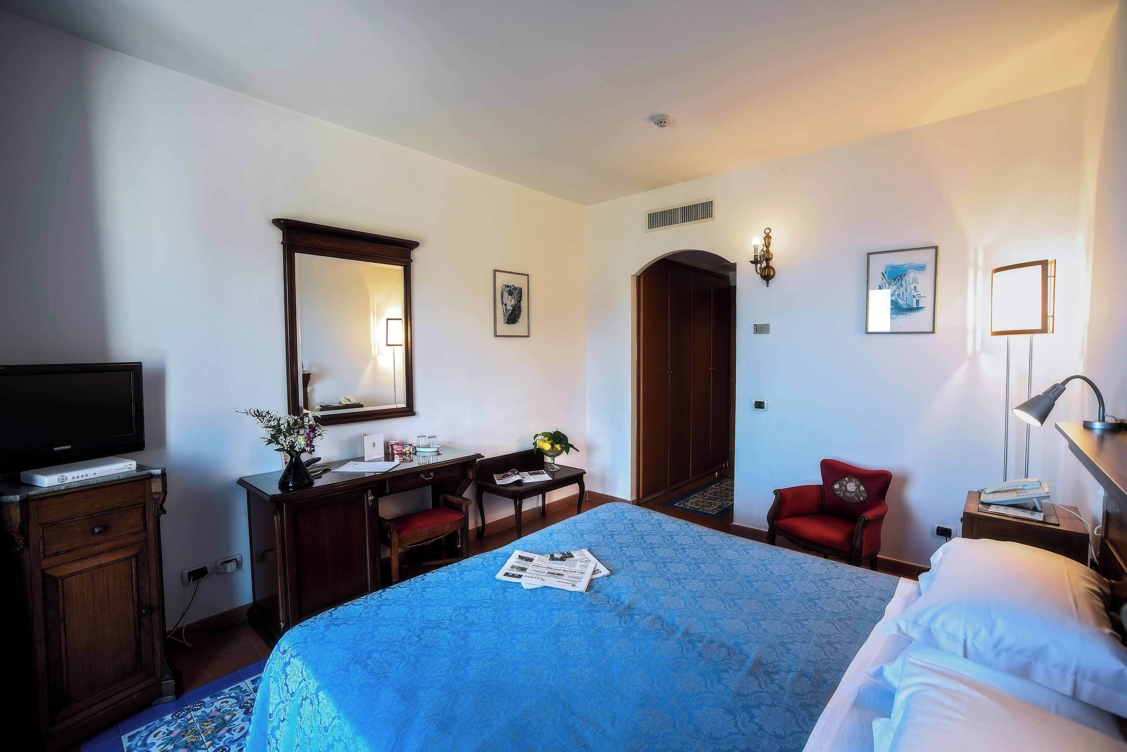 Een hotelkamer met een bed, een bureau met stoel, een televisie, een spiegel en een deur naar een andere kamer.