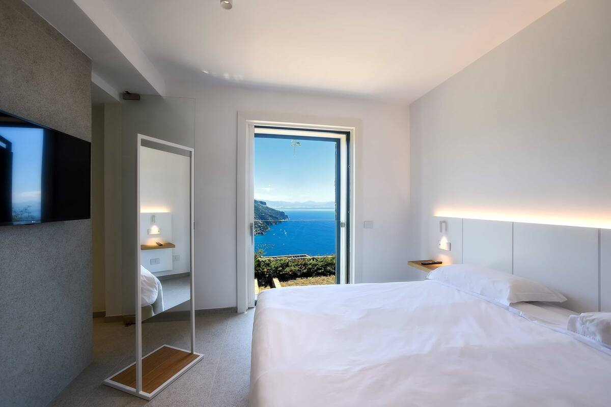 A modern hotel room with a large bed, a television, a mirror, and a door leading to a balcony with a view of the sea.